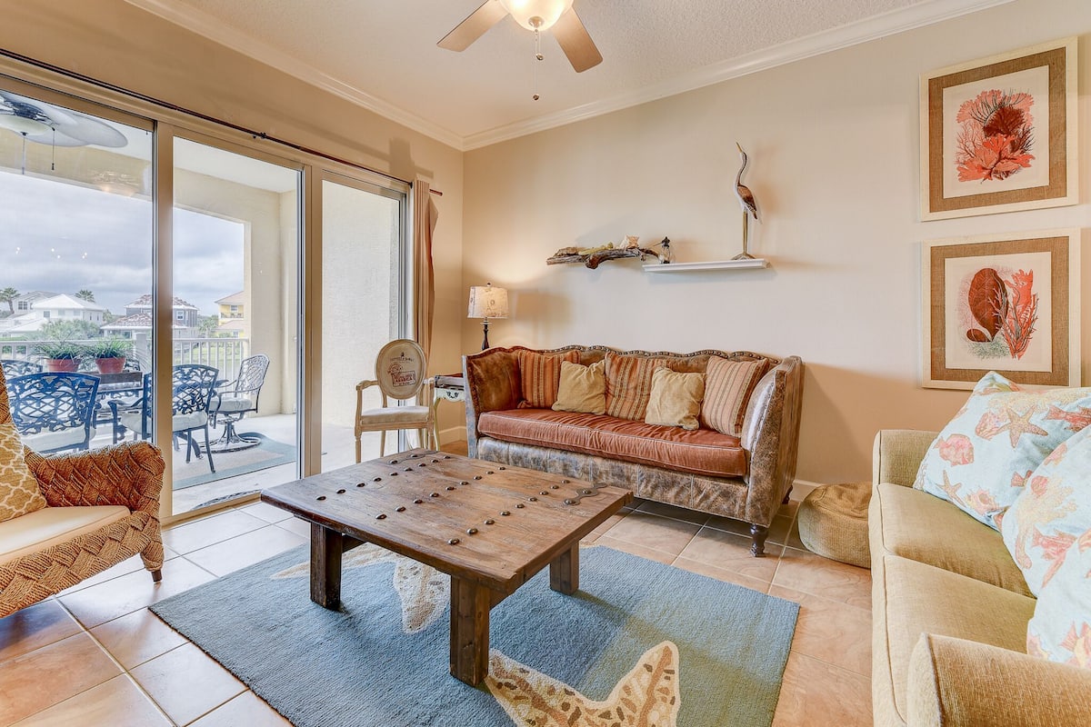 A spacious living room with a large window, a ceiling fan, a wooden coffee table, and a sofa with patterned cushions.