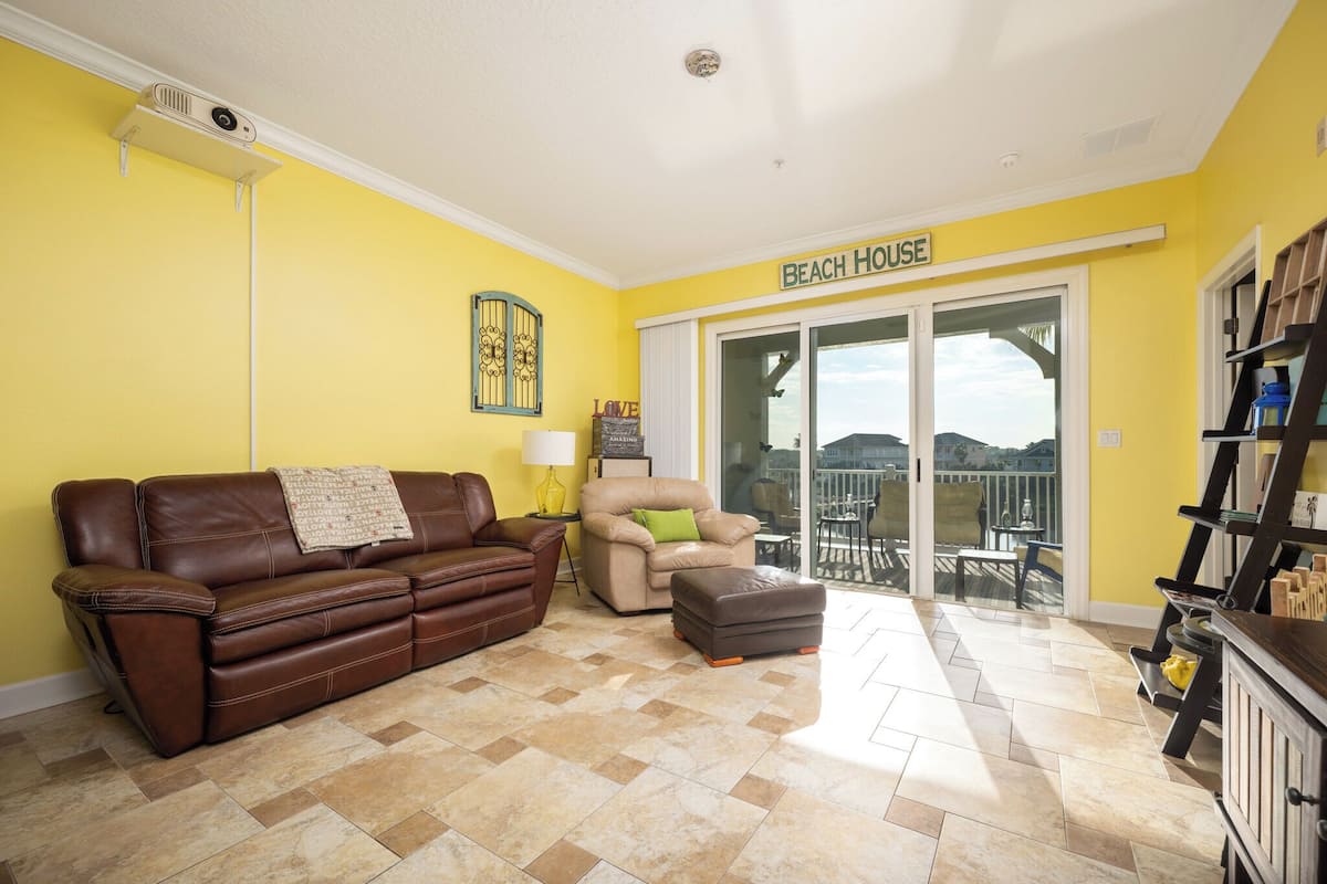 A living room with a brown leather sofa, a beige armchair, and a small ottoman. A sign above the door reads "BEACH HOUSE."