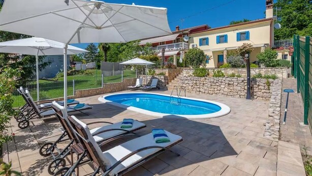 Une piscine entourée de chaises longues et de parasols, le tout délimité par un mur de pierre et bordé d’un bâtiment doté d’un balcon.