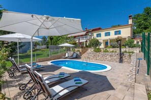 Une piscine entourée de chaises longues et de parasols, le tout délimité par un mur de pierre et bordé d’un bâtiment doté d’un balcon.