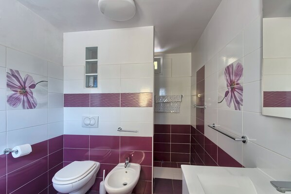 Une salle de bain équipée de toilettes, d’un bidet et d’un lavabo, avec des carreaux violets et blancs et un miroir.