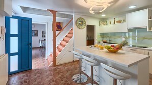 Une cuisine moderne avec des meubles blancs, un îlot central doté de tabourets, une horloge au mur et un escalier menant à une autre pièce.