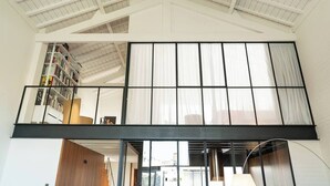 A high-ceilinged room with a mezzanine level, large windows, and a modern staircase.