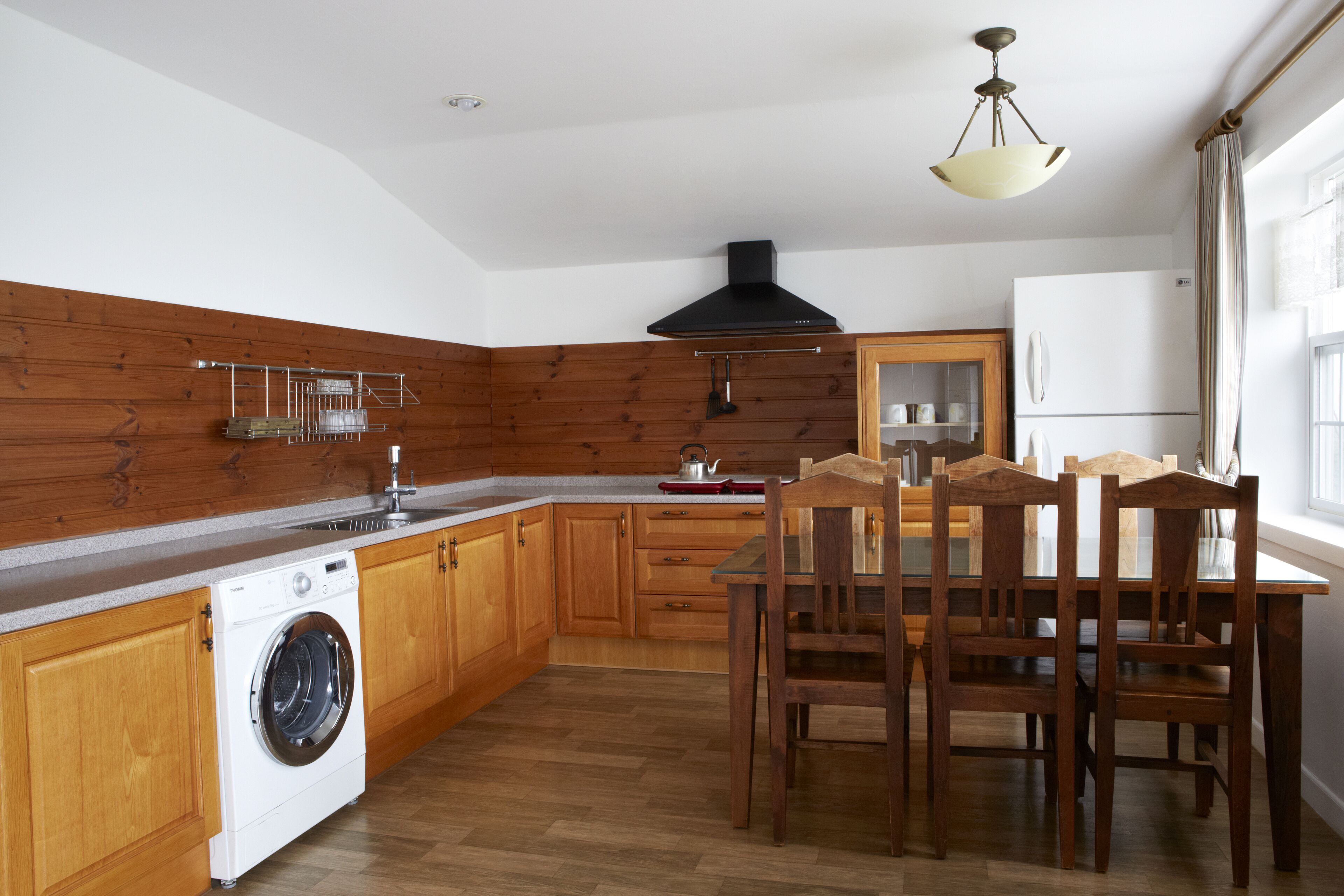A kitchen with wooden cabinets, a built-in washing machine, a dining table with chairs, and a hanging light fixture.