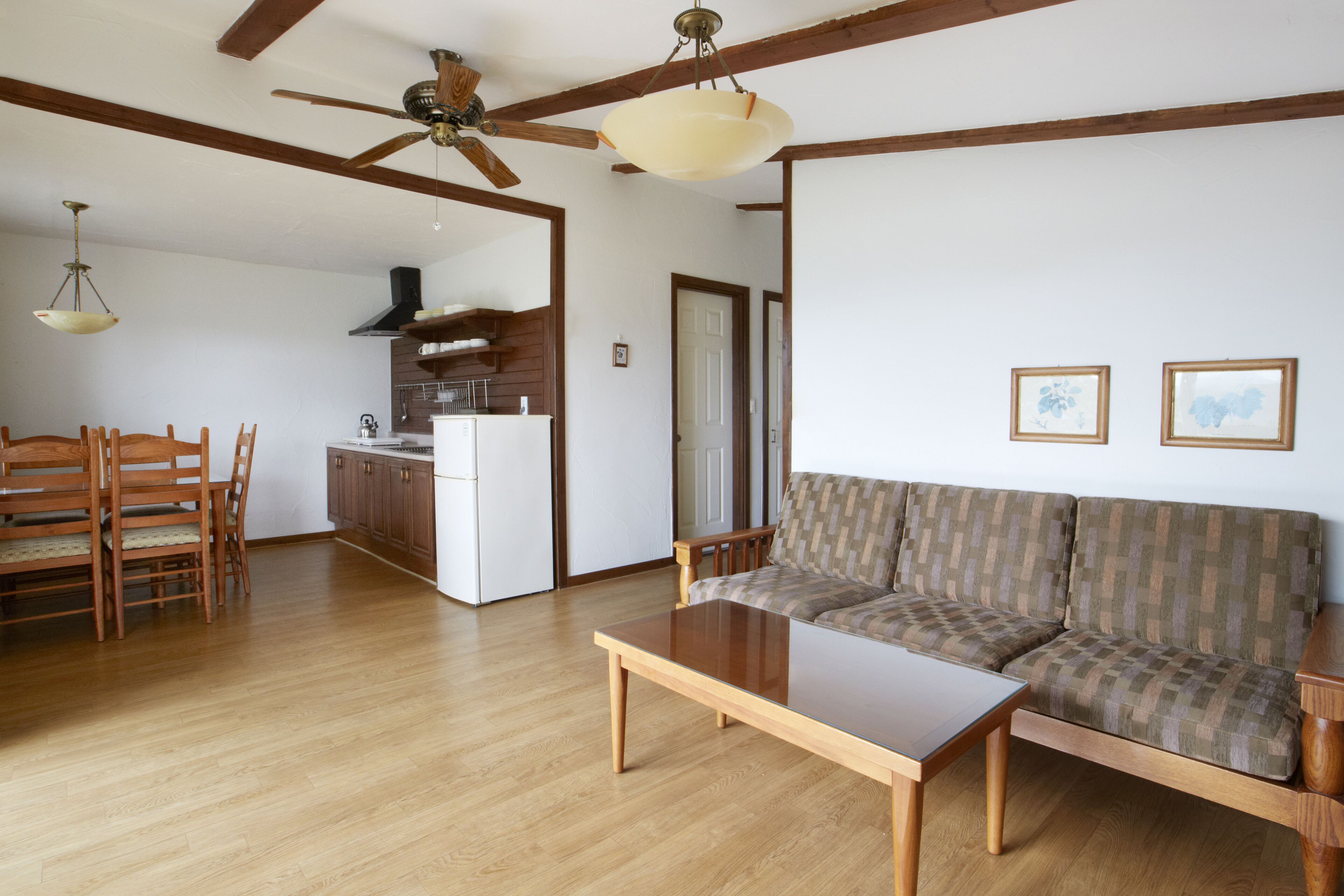 Amplia sala de estar con piso de madera, un sofá, una mesa de centro y una mesa de comedor con sillas. Se observa una cocina al fondo con un refrigerador y armarios.