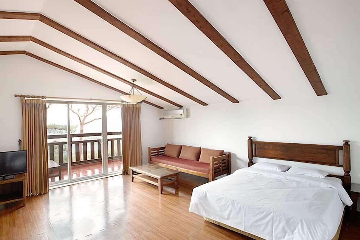 Quarto com teto de madeira, cama, sofá, televisão e varanda com cortinas.