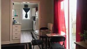 Une cuisine compacte équipée d’un réfrigérateur, d’un four à micro-ondes et d’une fenêtre avec des rideaux. On y trouve également un coin repas avec une table et des chaises, ainsi qu’un petit balcon agrémenté d’une plante en pot.