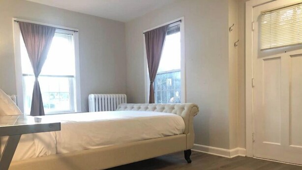 Une chambre à coucher avec un lit, un radiateur et deux fenêtres équipées de rideaux.