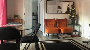 Un salon avec une table à manger en verre, deux chaises noires, deux fauteuils orange, un tapis noir et blanc, un petit sapin de Noël et un miroir au mur.