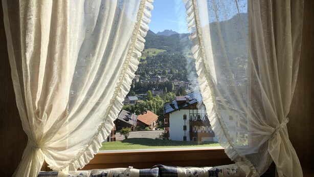 A window with sheer curtains, revealing a view of a town with houses and mountains in the background.