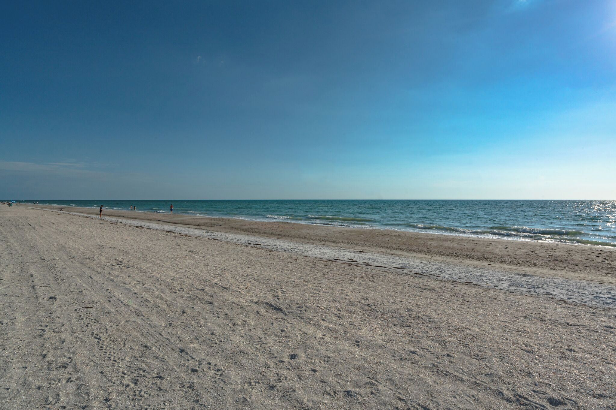Een breed zandstrand met zachte golven, een heldere lucht en enkele mensen in de verte.