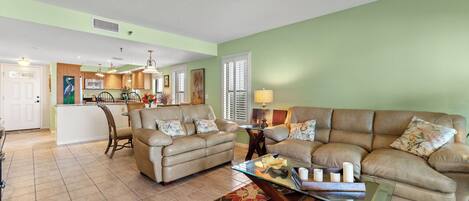 A spacious living room with a large beige sofa, a glass coffee table, and a patterned rug. A kitchen with wooden cabinets and a dining area with a chair and table are visible in the background.