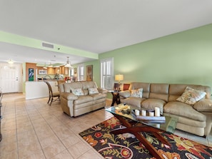 A spacious living room with a large beige sofa, a glass coffee table, and a patterned rug. A kitchen with wooden cabinets and a dining area with a chair and table are visible in the background.