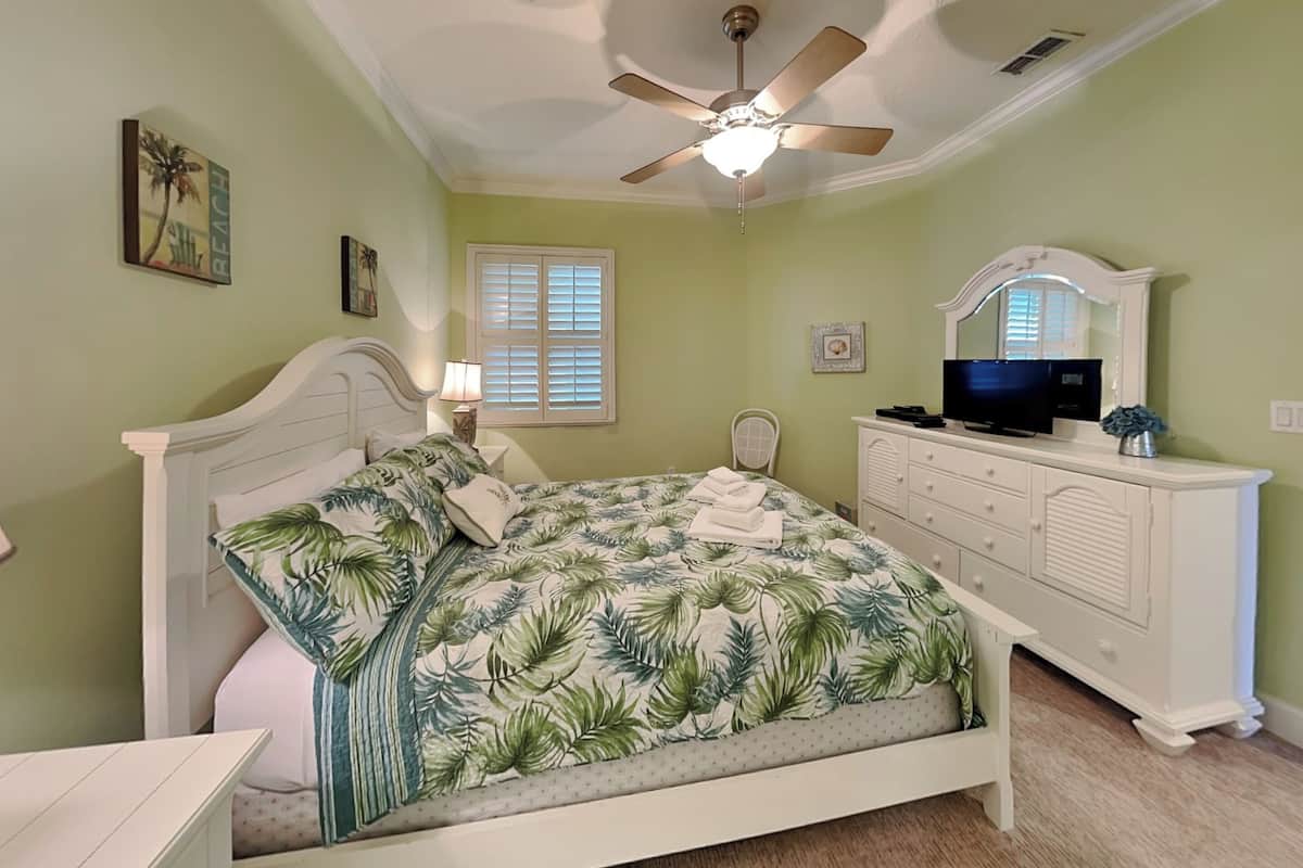 A bedroom with a bed, a dresser, a ceiling fan, a television, and a window with shutters.