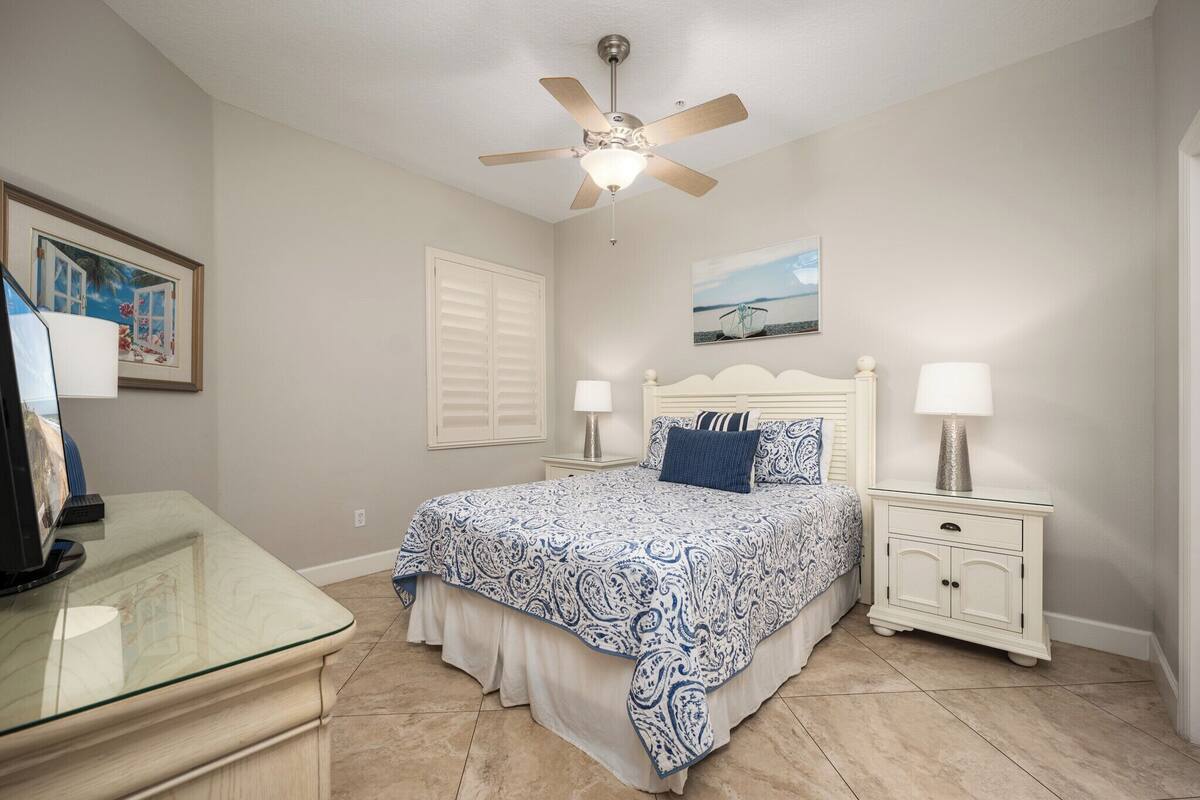 A bedroom with a bed, two bedside tables with lamps, a ceiling fan, a framed picture on the wall, and a glass-top desk with a computer monitor.
