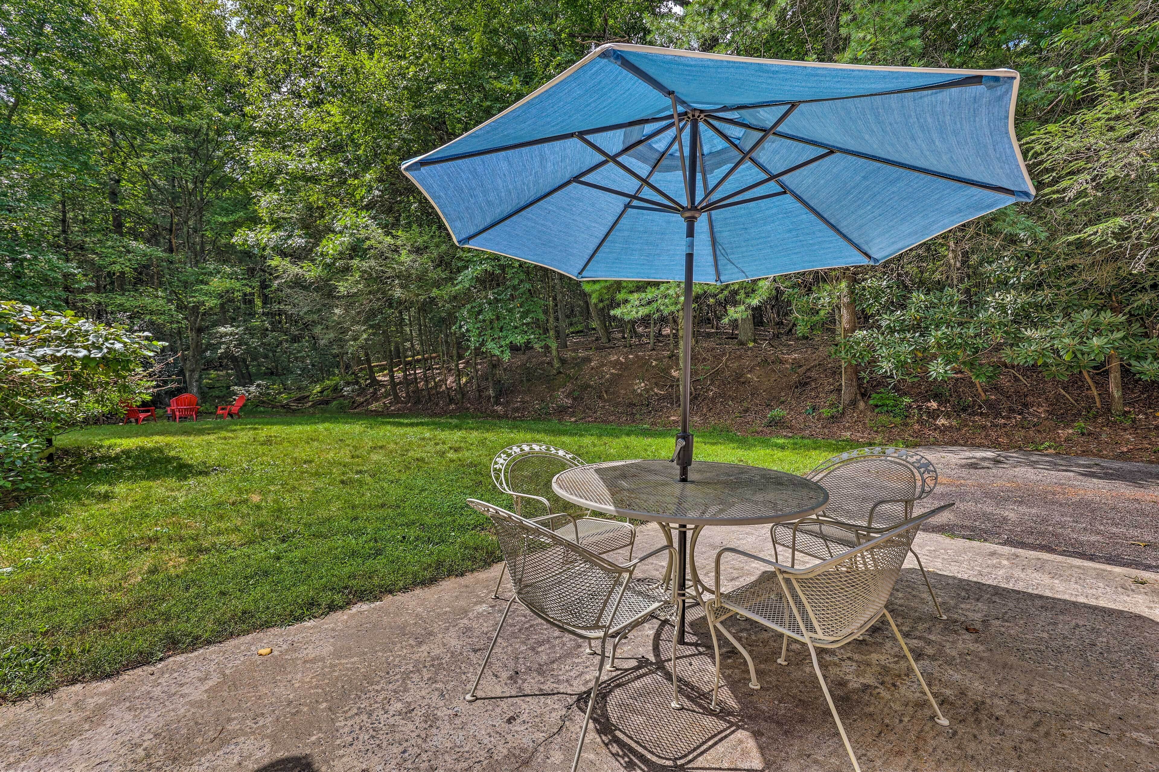 An outdoor patio with a round metal table and chairs, a blue umbrella, and a grassy area with trees in the background.