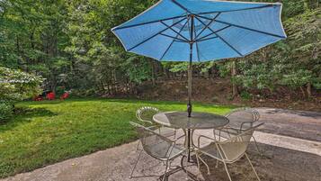 An outdoor patio with a round metal table and chairs, a blue umbrella, and a grassy area with trees in the background.