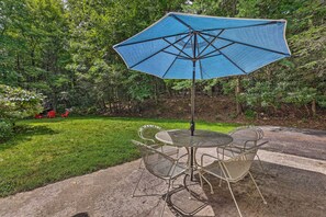 An outdoor patio with a round metal table and chairs, a blue umbrella, and a grassy area with trees in the background. - Near Hiking: Roaring Gap Retreat w/ Fire Pit! (Roaring Gap)