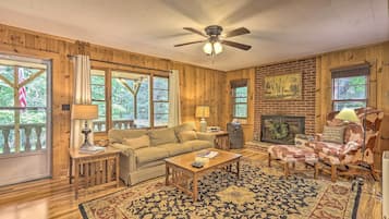 A cozy living room with wooden walls, a ceiling fan, a fireplace, and a large window overlooking greenery.