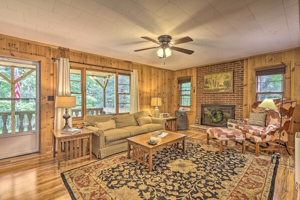 A cozy living room with wooden walls, a ceiling fan, a fireplace, and a large window overlooking greenery. - Near Hiking: Roaring Gap Retreat w/ Fire Pit! (Roaring Gap)
