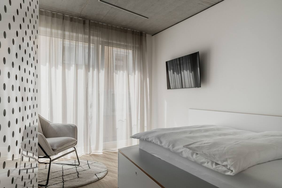 Un dormitorio moderno con cama, una silla y un ventanal con cortinas ligeras.