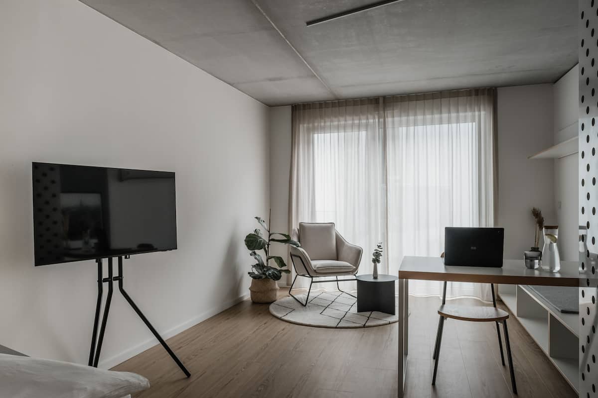 Una sala moderna con un televisor de pantalla plana sobre un soporte de trípode, una mesa redonda de madera, una computadora portátil, una silla y una planta en maceta.