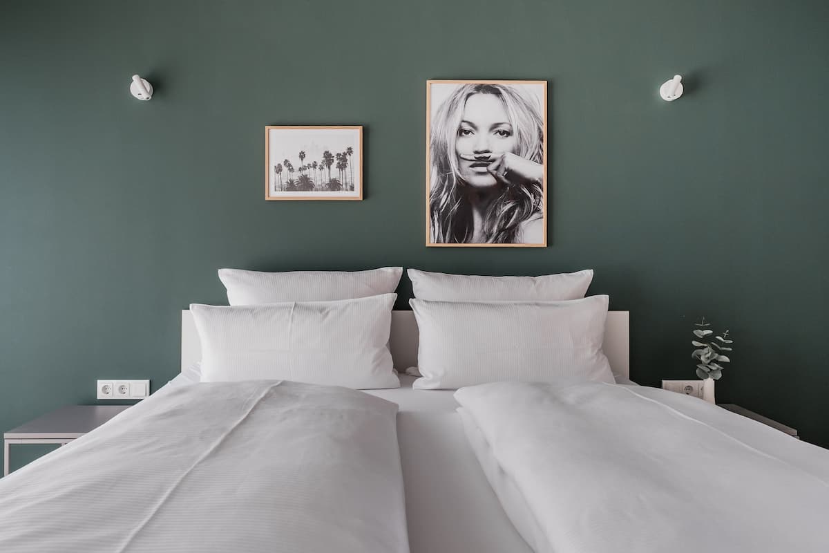 Un dormitorio con cama, dos almohadas, una mesita de noche, un cuadro enmarcado y una lámpara de pared.