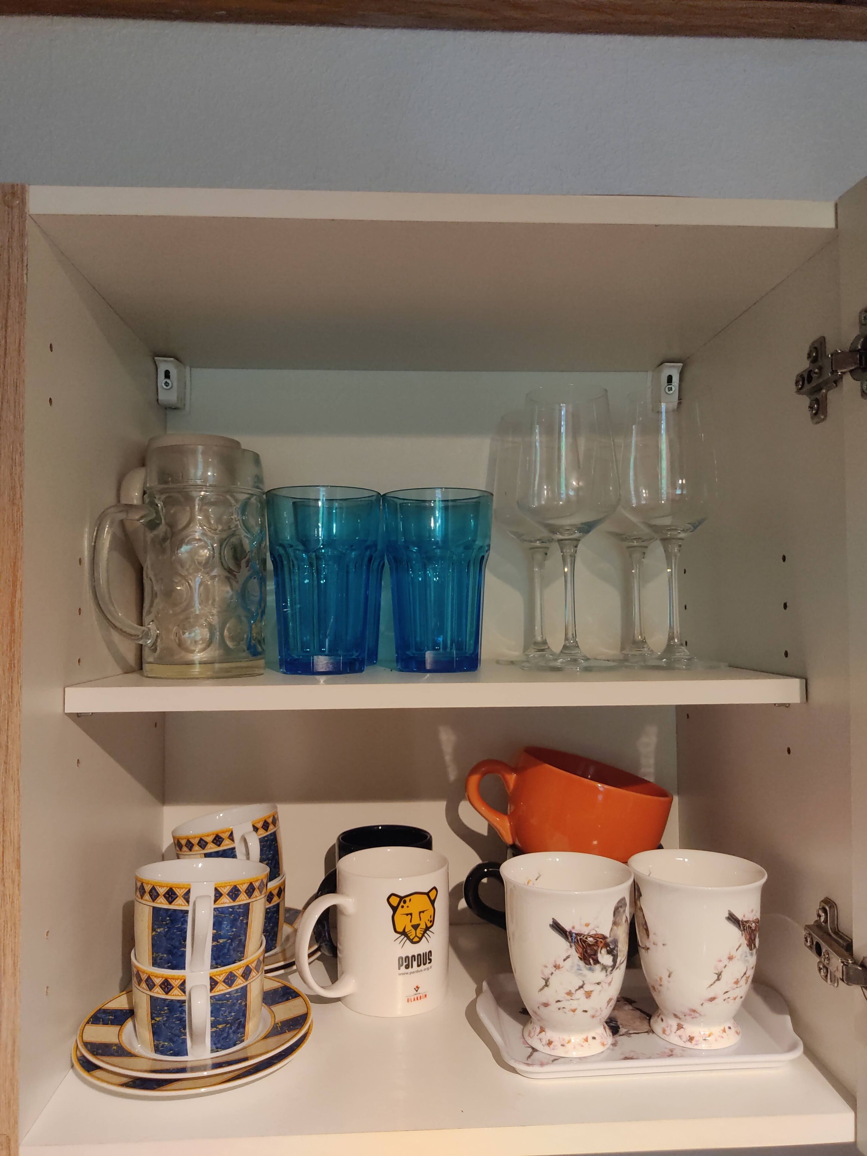 Uma estante com diversos copos, incluindo um jarro, copos azuis e taças de vinho. Abaixo, encontram-se canecas decorativas e uma chaleira.