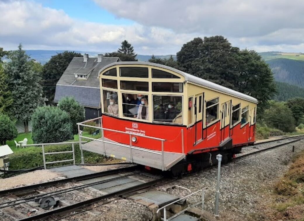 A red and yellow train with passengers, traveling on tracks surrounded by greenery and hills.