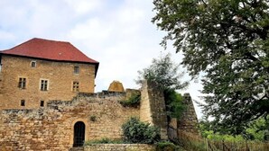 Un edificio histórico de piedra con techo rojo, un arco central y un pequeño patio con plantas en macetas y mobiliario exterior.