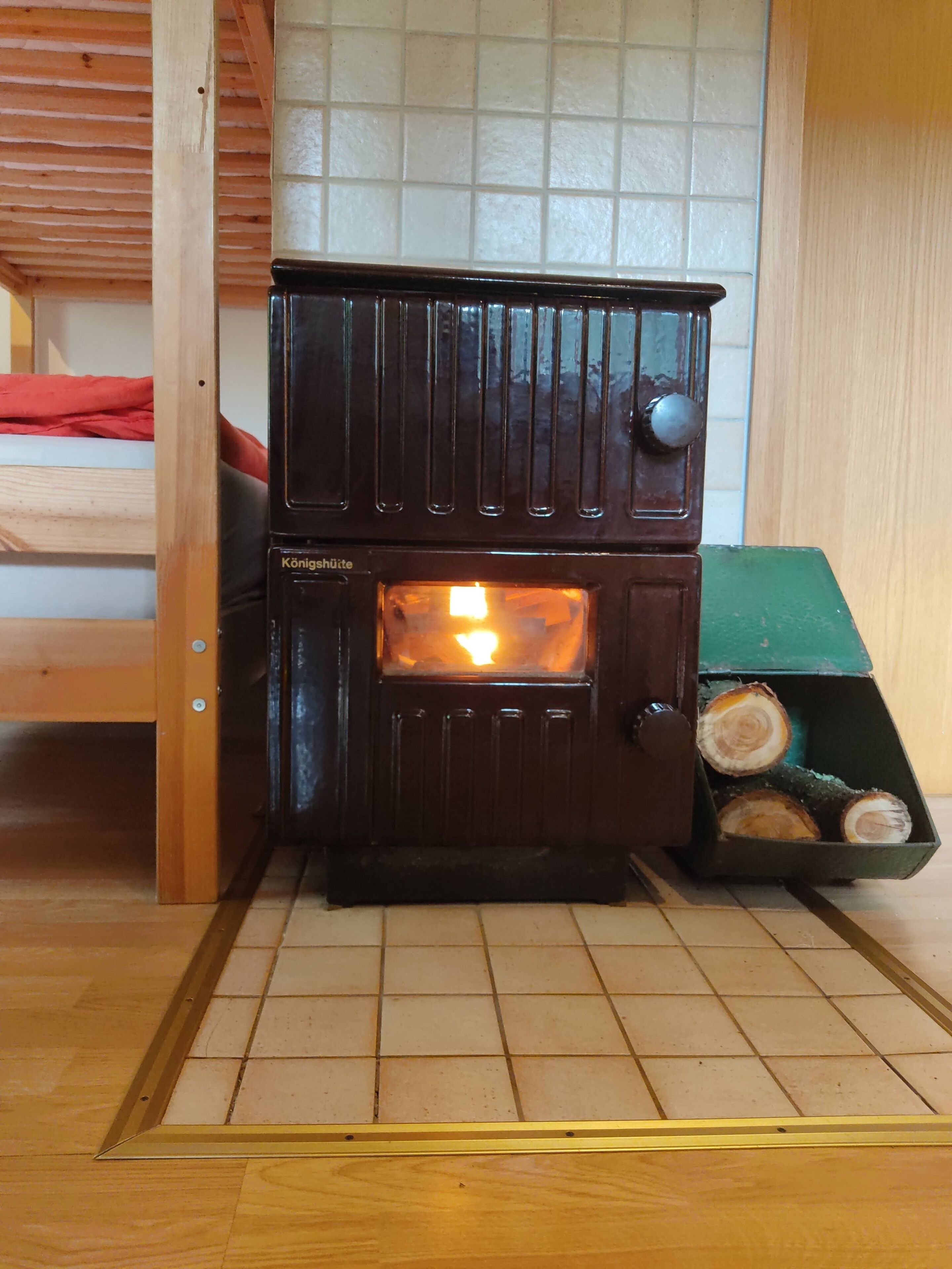 A small, two-tiered wood-burning stove with a visible fire, placed in a room with wooden flooring and a tiled wall.