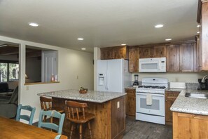 A kitchen with wooden cabinets, a refrigerator, a microwave, a stove, and a granite countertop island with two chairs.