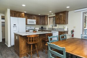 A kitchen with wooden cabinets, a white refrigerator, a microwave, a granite countertop, and a dining table with chairs.