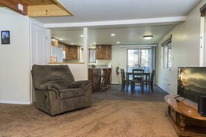 Una sala de estar con un sillón reclinable, un comedor con mesa y sillas, y una cocina con armarios de madera y un refrigerador.