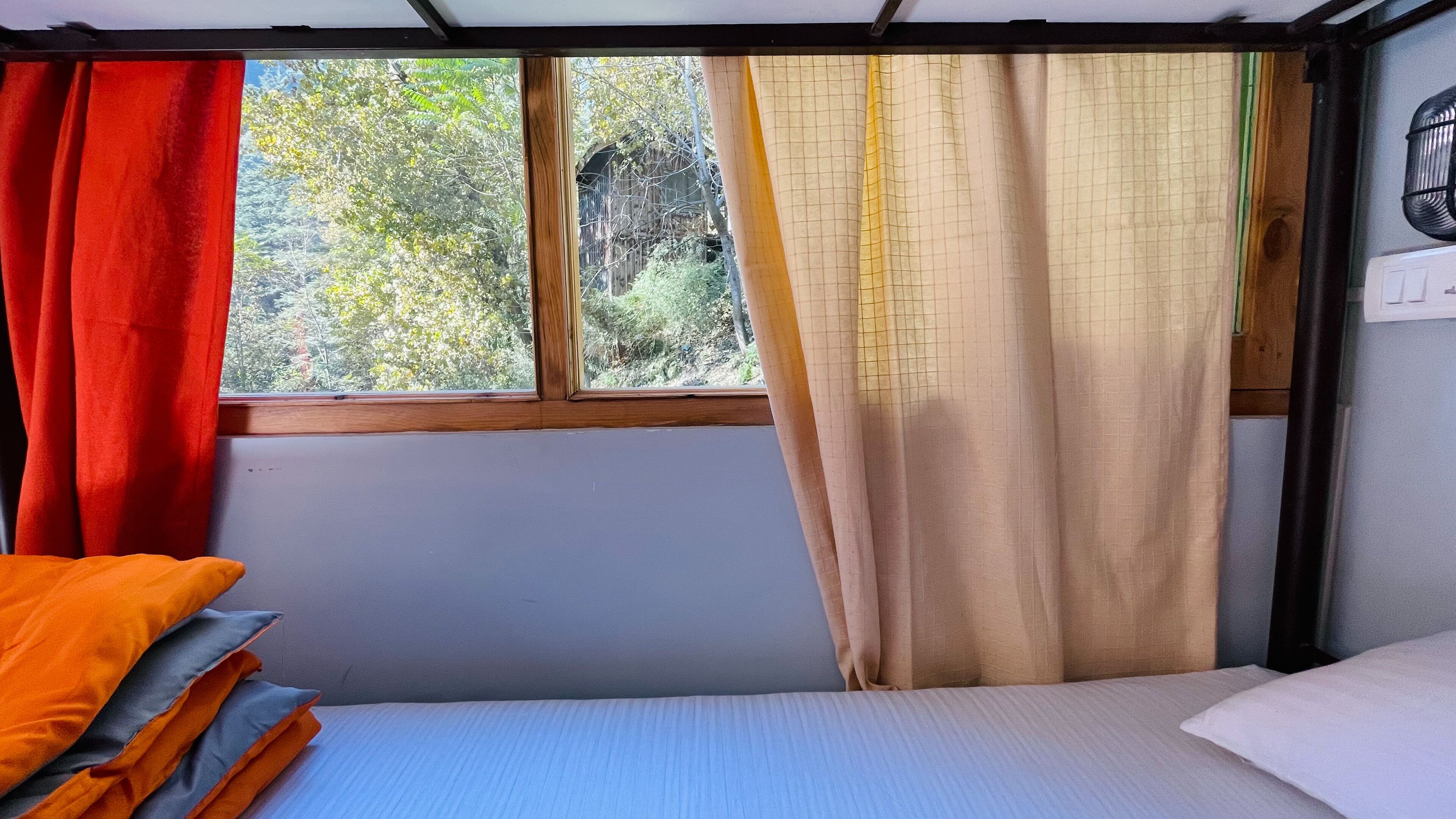 A single bed with a blue mattress and white pillows, a window with a view of trees, and a red curtain on the left.