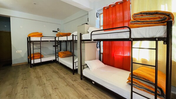 A dormitory room with bunk beds, a small cabinet, and a window with orange curtains. - Madpackers Jibhi - Hostel (Banjar)