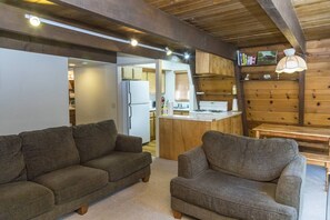 A cozy living area with two sofas, a wooden ceiling, and a kitchen in the background.
