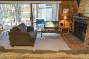 A cozy living room with a sofa, TV, fireplace, and a view of a deck and trees.