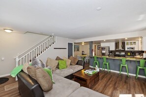 A modern living room with a sofa, coffee table, and a dining area with chairs. - Walking Distance to Snow Summit Ski Resort!! Beautifully Updated 3 BED 2 BATH! Wood Burning Fireplace! Smart TVs w/ Streaming Services! (Big Bear Lake)