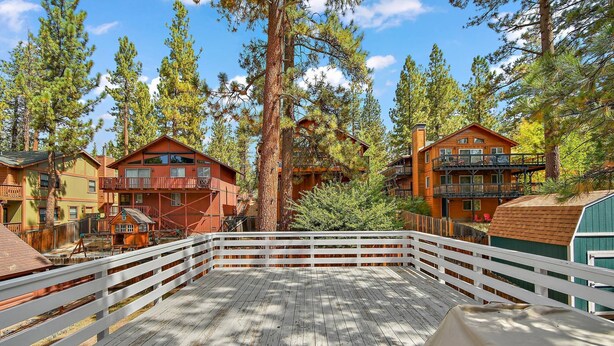 A wooden deck with a white railing, surrounded by tall pine trees and cabins with balconies.