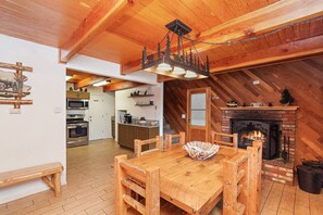 A wooden dining table with chairs, a bench, a fireplace, and a kitchen in the background.
