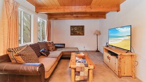 A cozy living room with a wooden ceiling, a large window, a sofa with patterned cushions, a coffee table, and a television displaying a beach scene.