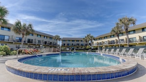 Una piscina circular con indicador de profundidad de 1 metro 18 centímetros, rodeada de sillas de descanso y palmeras, y un edificio de varios pisos al fondo.