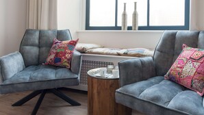 Een kamer met twee grijze fauteuils, een klein houten bijzettafeltje, een raam met uitzicht en een radiator.