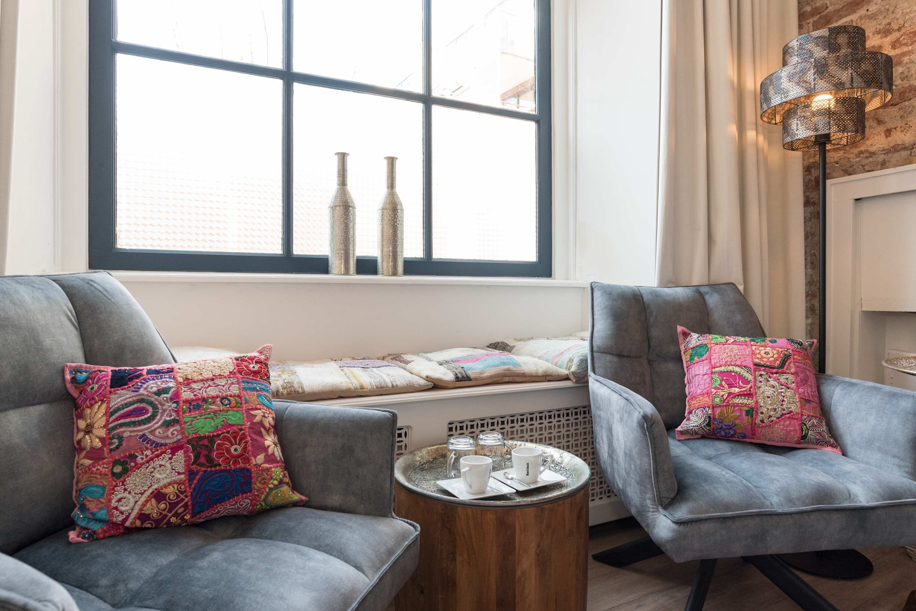 A room with two grey armchairs, a small wooden side table with cups and a tray, and a window with decorative bottles.