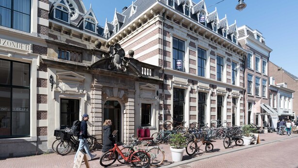 Een historisch gebouw met een rij fietsen voor de deur, een persoon die loopt en een bordes op de stoep.