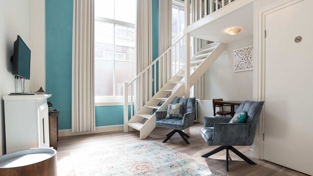 Un salon moderne avec un escalier, deux fauteuils, un téléviseur à écran plat et un tapis décoratif.