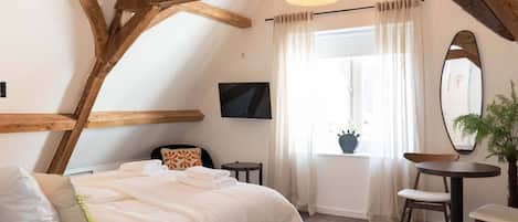 Een slaapkamer met een bed, een televisie, een raam met gordijnen, een spiegel en een klein tafeltje met stoelen.