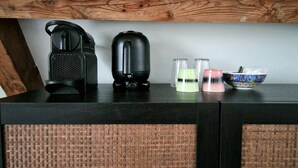 Een zwarte koffiemachine, een zwarte elektrische waterkoker, twee glazen, een roze kaars en een schaal op een donker houten tafel.
