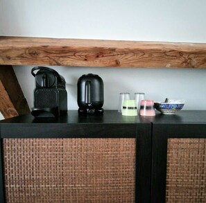Une petite cuisine équipée d’une machine à café, d’une bouilloire, de deux verres et d’un bol, le tout posé sur un plan de travail de couleur sombre.